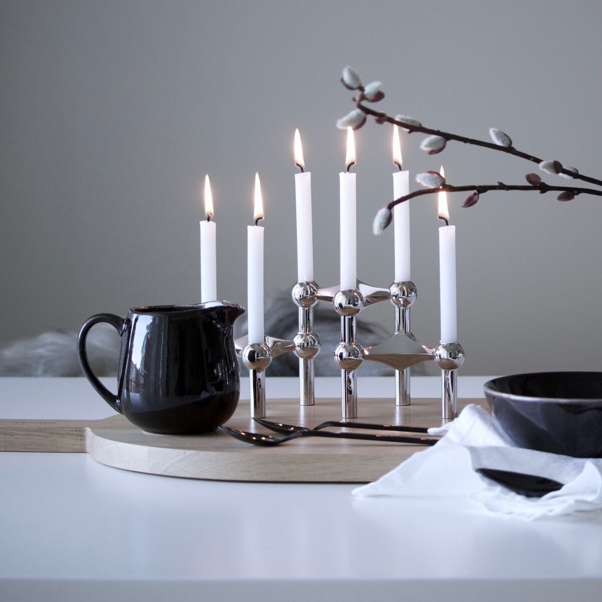 Ein moderner Kerzenständer aus Chrom steht auf einem weißen Tisch. Darauf befinden sich mehrere weiße Kerzen, die brennen. Neben dem Kerzenständer steht eine schwarze Kanne und eine schwarze Schüssel auf einem Holzbrett. Ein Ast mit dekorativen Blättern ragt in das Bild hinein.