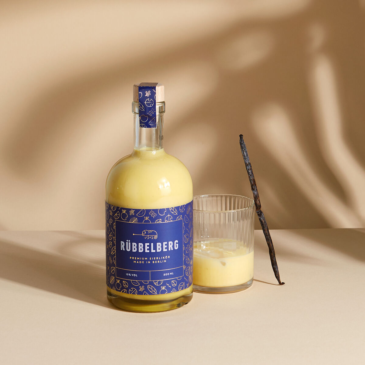 Eine elegante Glasflasche mit einem blauen Etikett des Premium Eierlikörs von Rübbelberg, daneben ein Glas mit einem Vanillestab, auf einem beige Hintergrund fotografiert.