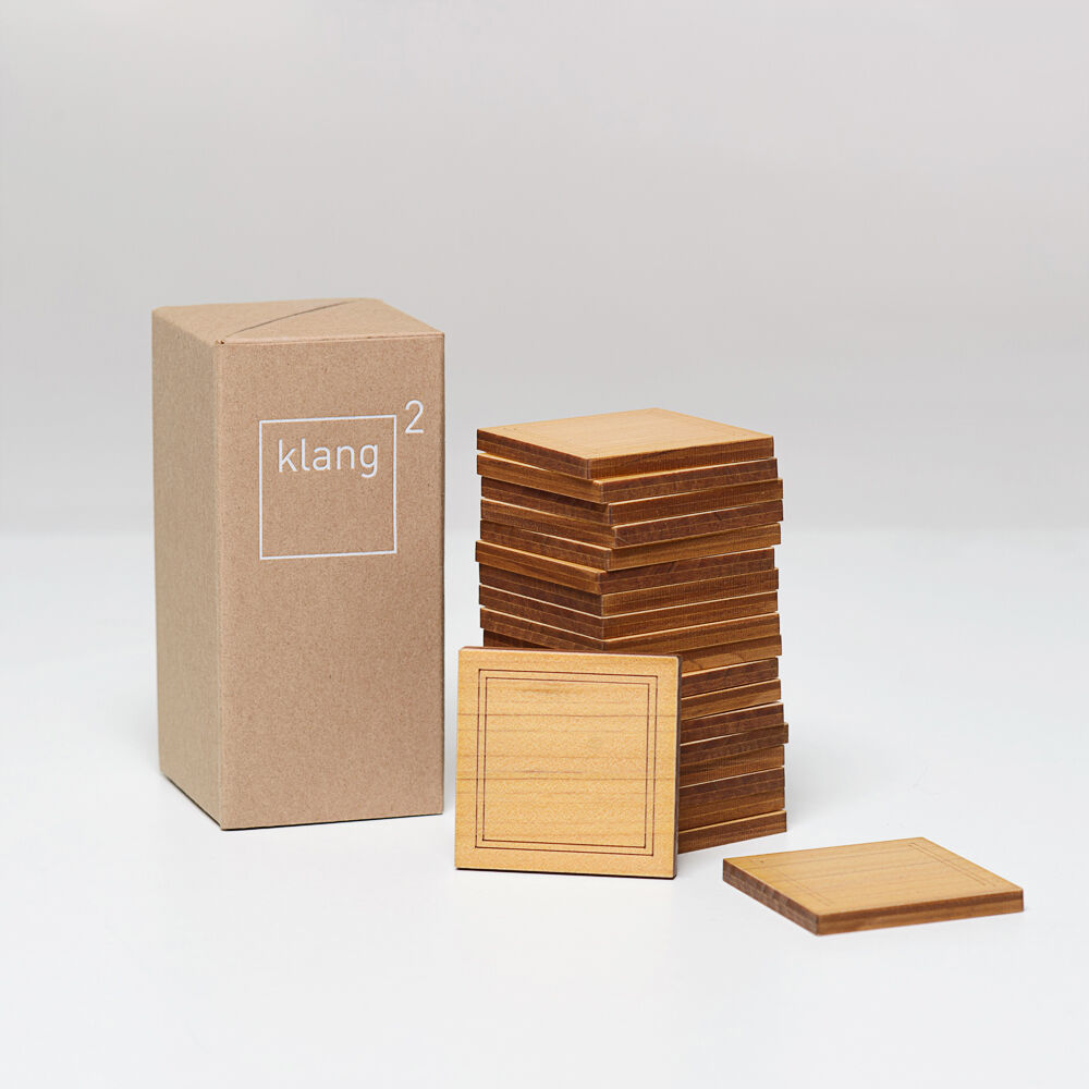 Klang² Spielesammlung 'Geigenbauer' bestehend aus hölzernen quadratischen Spielsteinen, gestapelt neben einer Kartonverpackung. Das minimalistische Design und die natürlichen Holzfarben dominieren das Bild. Ideal für Kinder und Erwachsene zur Förderung von Kreativität und handwerklichen Fähigkeiten.
