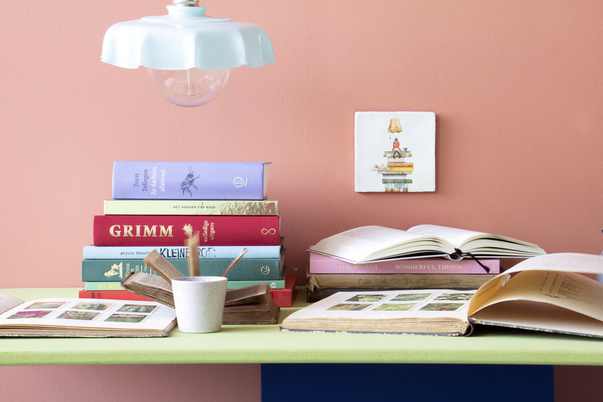 Eine Szene mit einem Bücherstapel auf einem Schreibtisch. Im Hintergrund ist eine Keramikfliese mit einem Bild von einem Bücherwurm platziert. Die Wände sind rosa, und es gibt ein helles, hängendes Deckenlicht. Auf dem Schreibtisch liegen mehrere offene Bücher und eine Tasse.