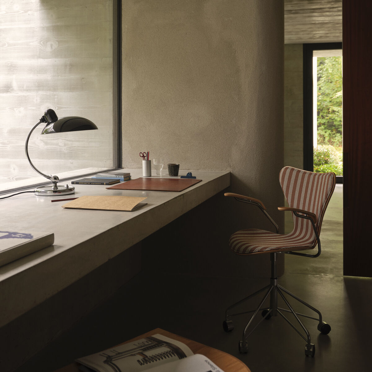 Schreibtischstuhl "Serie 7" von Fritz Hansen steht an einem modernen, minimalistischen Schreibtisch vor einem großen Fenster. Der Raum wirkt hell und offen, die Stuhlpolsterung ist gestreift. Auf dem Tisch liegen Schreibtischutensilien und eine schwarze Lampe.