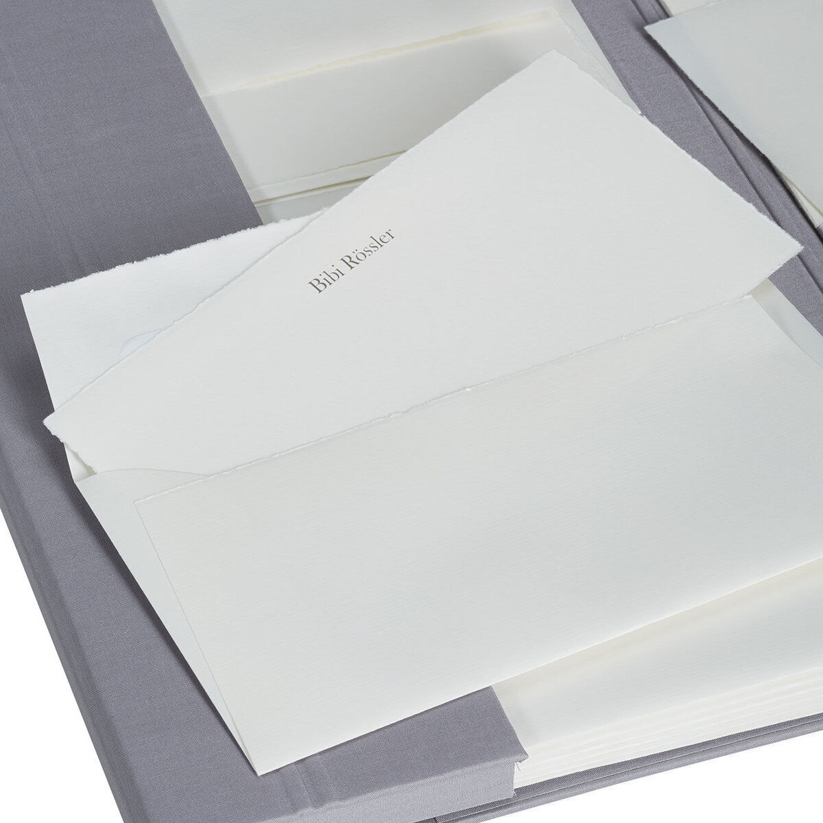 Ein elegantes Set aus handgeschöpftem Büttenpapier in einer hochwertigen Briefpapiermappe. Die Mappe ist in einem neutralen Grauton gehalten und enthält mehrere Blätter und Umschläge in Weiß und Creme. Das Produkt vermittelt einen Hauch von Luxus und Eleganz, ideal für besondere Anlässe oder als exquisites Geschenk.