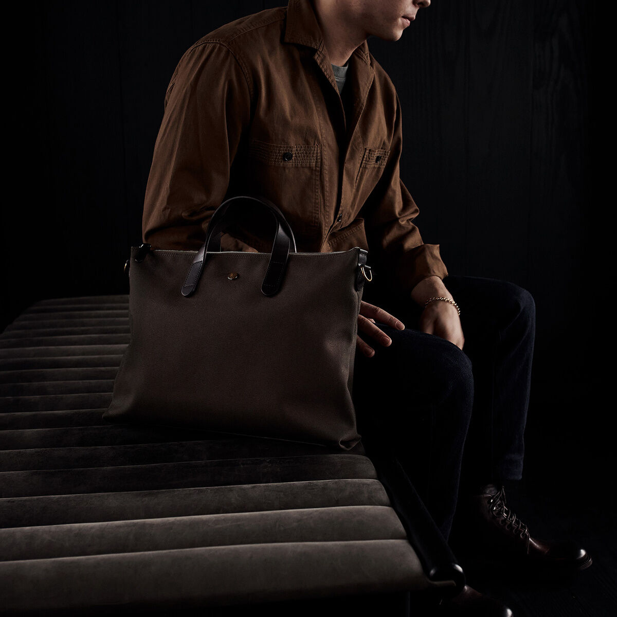 Ein Mann sitzt in einem dunklen Raum und hält eine elegante braune Laptop-Tasche aus Leder. Die Szene ist minimalistisch und strahlt Eleganz aus.