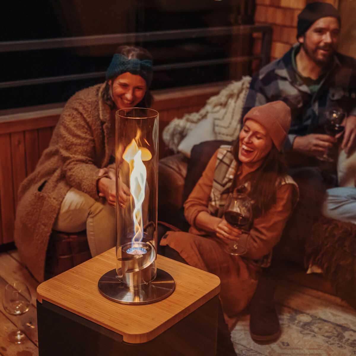 Eine Gruppe von Freunden sitzt im gemütlich beleuchteten Innenbereich auf einer Holzcouch um ein Tischfeuer SPIN 1200, das in einem Glaszylinder brennt. Sie genießen eine entspannte Atmosphäre.