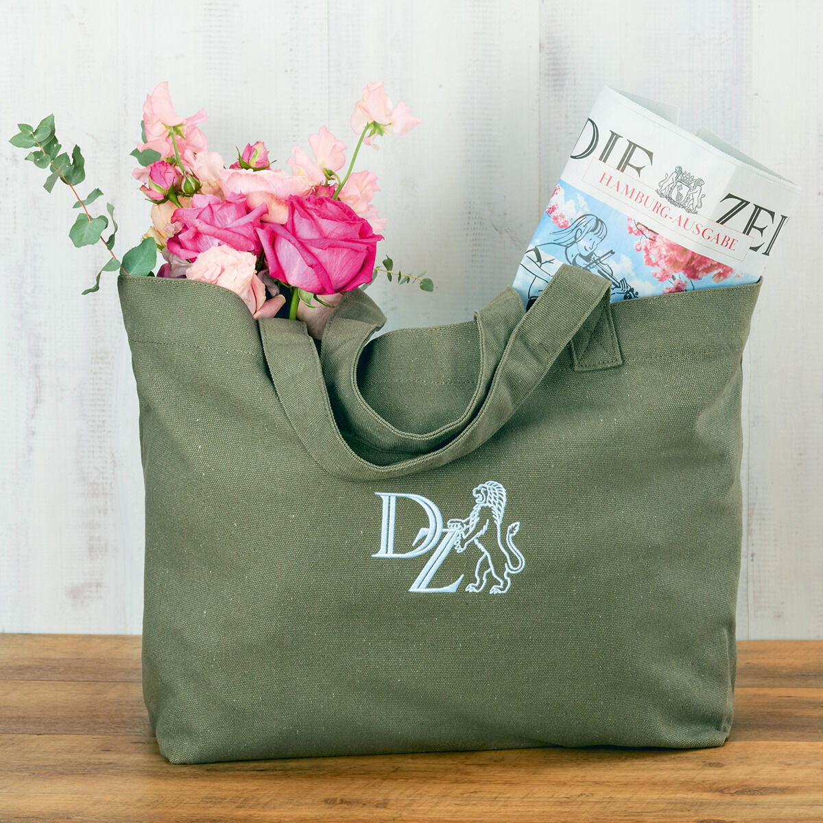 Grüne Canvas-Tasche von ZEIT, gefüllt mit rosa Blumen und einer Zeitung, auf einem Holztisch vor einer hellen Wand.