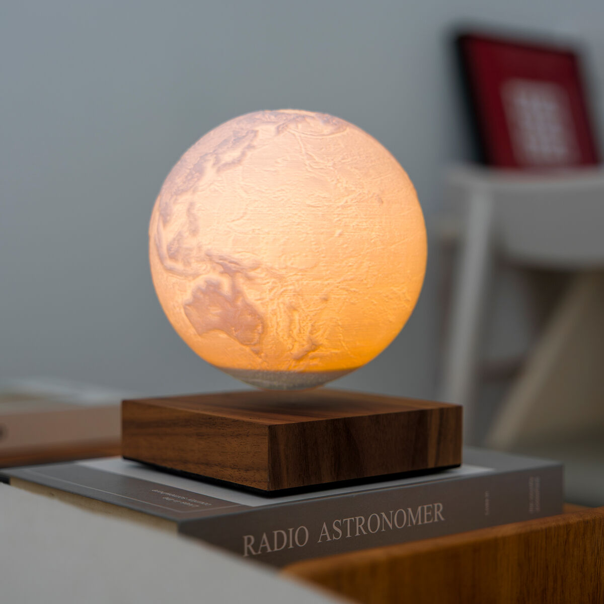 Die Leuchte »Smart Earth Lamp« von Gingko Design Ltd zeigt eine schwebende, leuchtende Weltkugel auf einem rechteckigen Holzsockel. Das Licht erzeugt einen warmen gelblichen Farbton. Im Hintergrund sind Bücher und Möbelstücke in einem modernen, dezenten Raum zu erkennen.