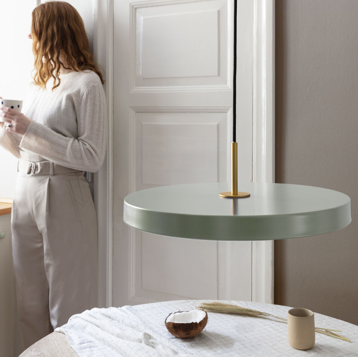 Eine moderne Pendelleuchte namens Asteria Medium hängt in einem stilvoll eingerichteten Raum. Eine Frau in hellen Kleidern steht am Fenster und trinkt aus einer Tasse. Der Hintergrund zeigt neutrale Farbtöne und minimalistische Dekorationselemente.