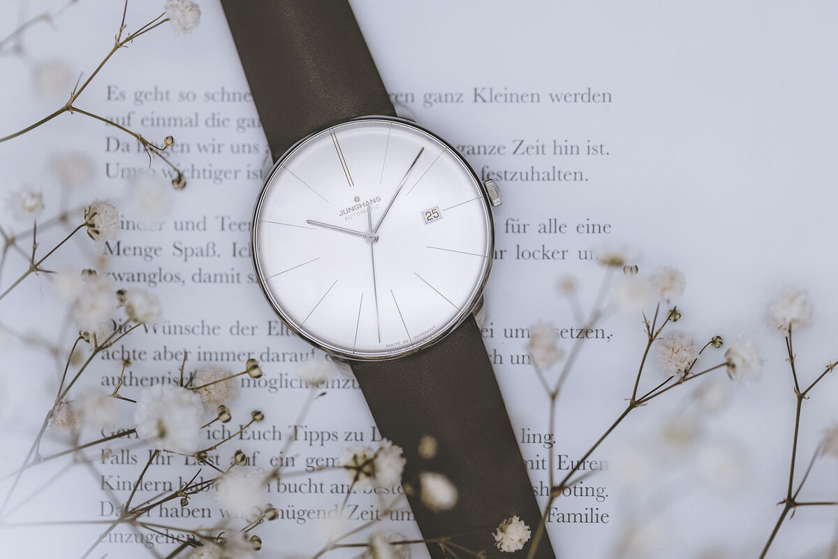 Eine elegante Armbanduhr der Marke Junghans, Modell ZEIT-Sonderedition »Meister fein Automatic«. Die Uhr besitzt ein minimalistisches Design mit einem weißen Zifferblatt und filigranen silbernen Zeigern. Das Armband ist schwarz. Im Hintergrund sind zarte weiße Blüten und ein Buch mit Text zu sehen.