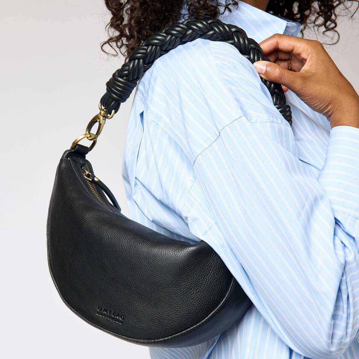 Frau trägt eine elegante, halbmondförmige schwarze Tasche mit geflochtenem Schulterriemen über der Schulter. Die Tasche hat goldene Elemente und passt zu einem hellblauen Oberteil.