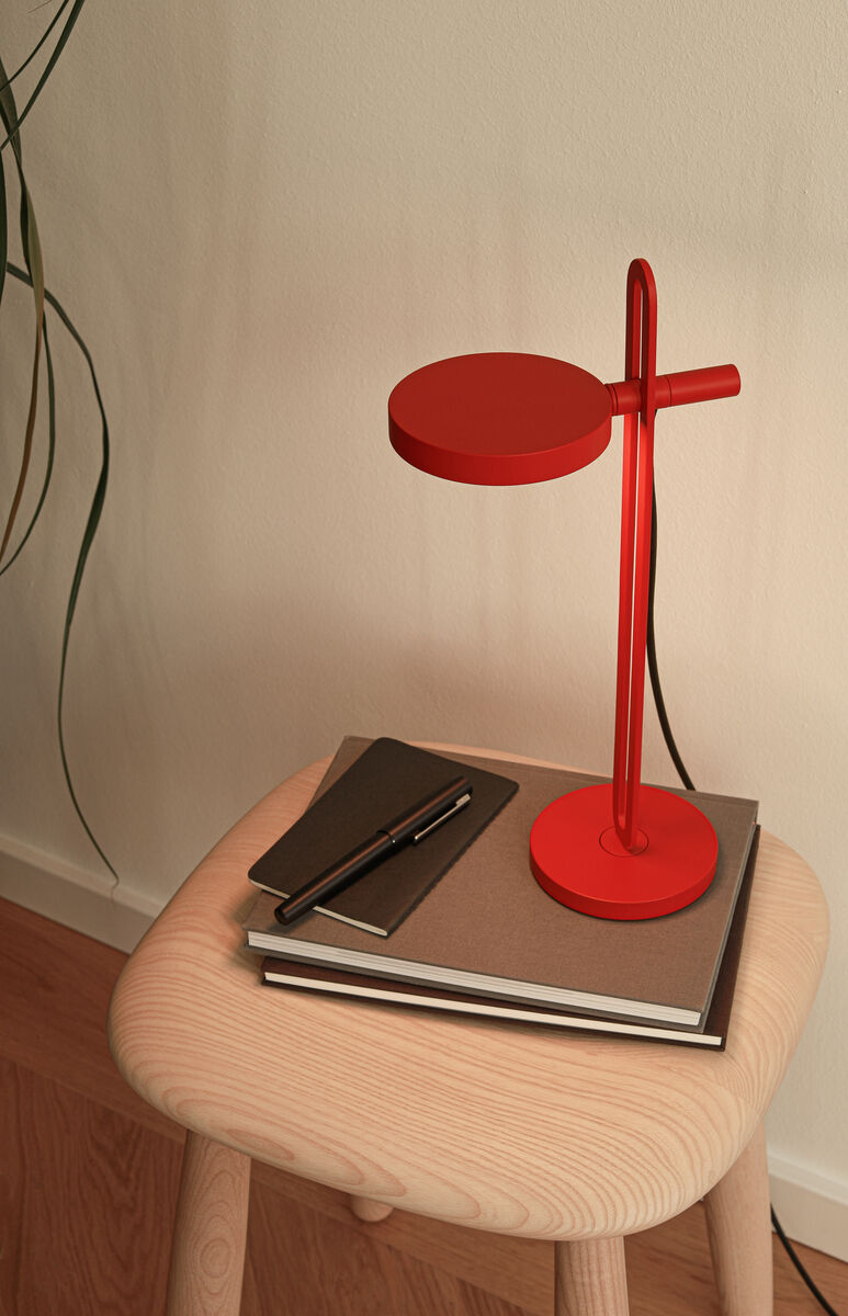 Eine rote Tischlampe steht auf einem hellen Holzhocker, darunter liegen zwei geschlossene Bücher und ein schwarzes Notizbuch mit einem Stift darauf. Im Hintergrund erkennt man eine hellbeige Wand mit einer dünnen, überhängenden Pflanze.