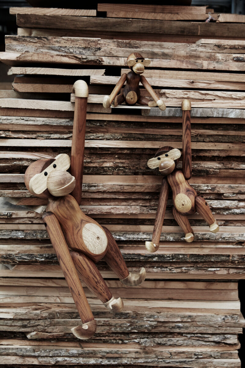 Drei Holzfiguren in Form von Affen, entworfen von Kay Bojesen, hängen und sitzen auf einem Stapel Holzbretter. Sie bestehen aus unterschiedlichen Holztönen und sind handgefertigt.