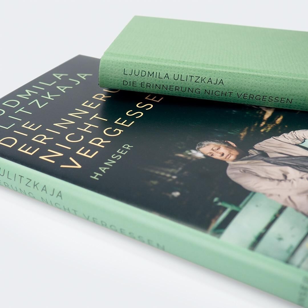 Das Bild zeigt zwei Ausgaben des Buches 'Erinnerung nicht vergessen' von Ljudmila Ulitzkaja, veröffentlicht vom Hanser Verlag. Beide Bücher haben grüne Umschläge, wobei eines flach auf dem Hintergrund liegt und das andere aufgerichtet ist. Auf dem flachen Buch ist das Bild einer älteren Person zu sehen.