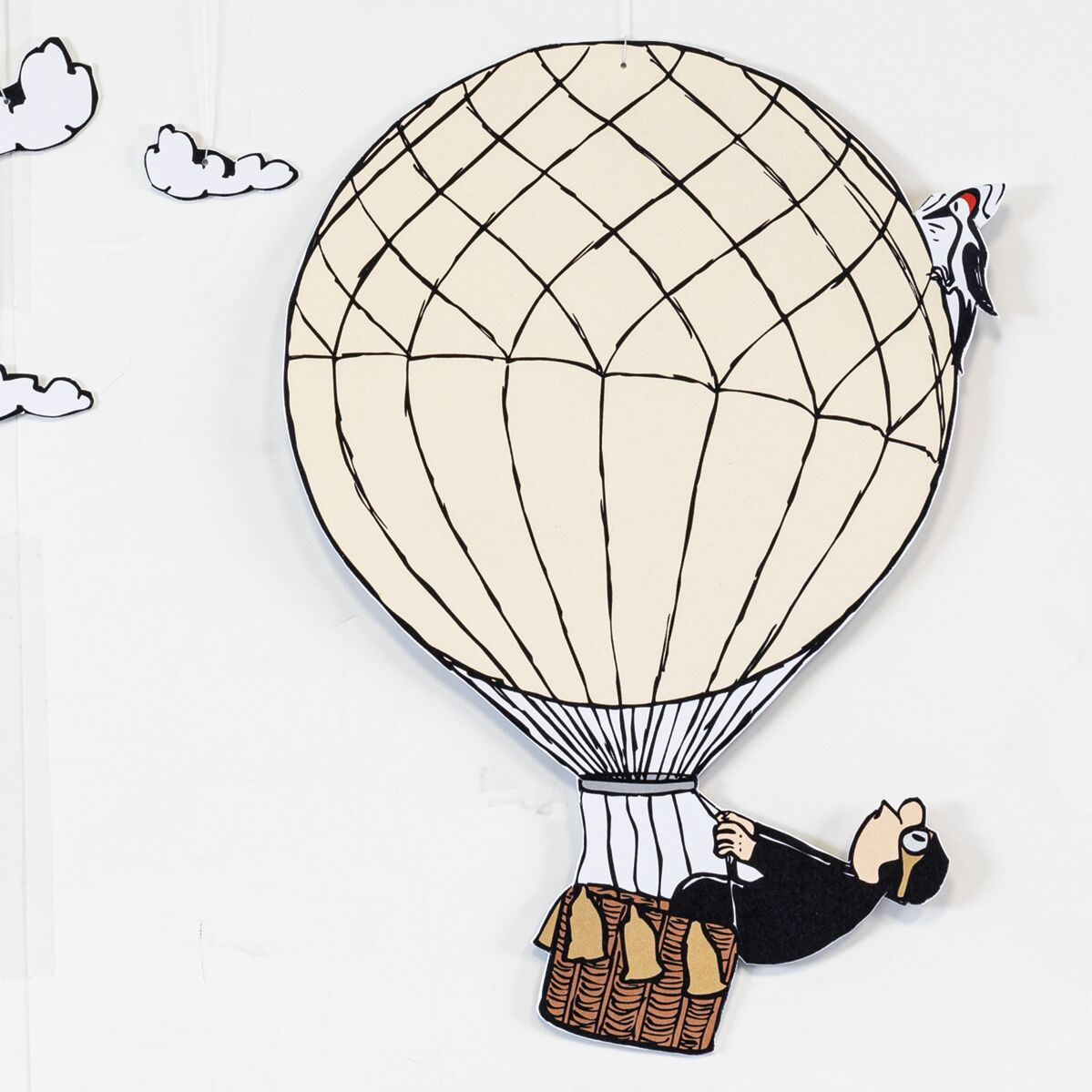 Das Bild zeigt das Decken-Mobilé "Der Ballonfahrer" von Loriot, herausgegeben von Ars mundi. Ein gezeichneter Herr im schwarzen Anzug sitzt in einem braunen Heißluftballon-Korb, am Ballon hängt ein Comic-Vogel. Der Ballon ist cremefarben mit schwarzen Linien, kleine Wolken schweben im Hintergrund.