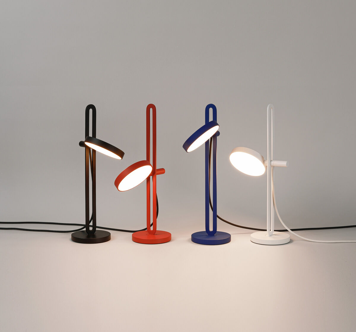 Vier Tischlampen des Modells »Echo« in verschiedenen Farben (schwarz, rot, blau und weiß) stehen nebeneinander auf einem Tisch. Jede Lampe besitzt ein minimalistisches Design mit einem runden Fuß und einem schlanken, aufrechten Ständer, der in einen schwenkbaren Lampenschirm mündet.
