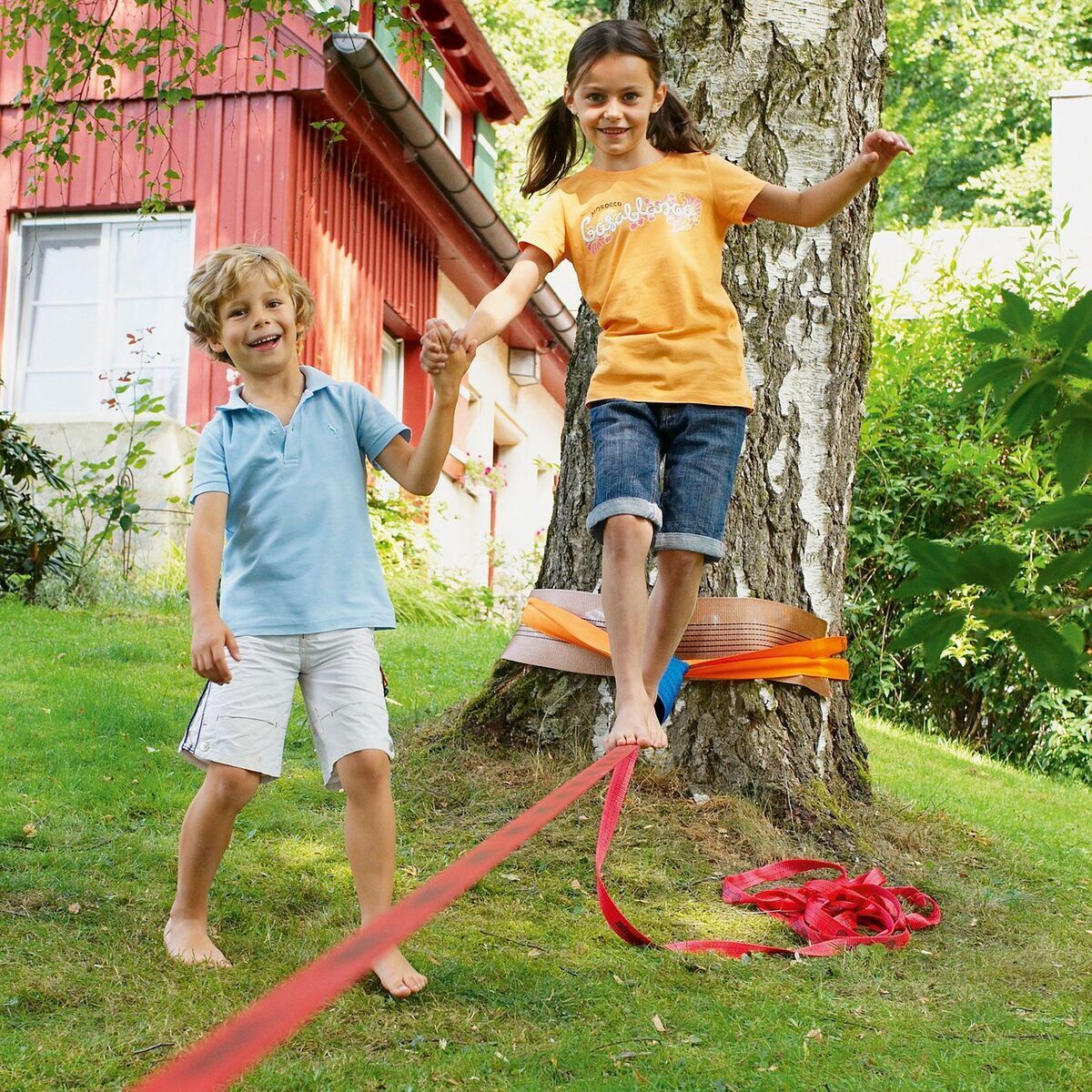 Zwei Kinder spielen auf einer blauen Slackline im Garten. Ein Junge in einem blauen T-Shirt und weiße Shorts hält die Hand eines Mädchens, das ein orangefarbenes T-Shirt und Jeans trägt. Die Slackline ist zwischen zwei Bäumen gespannt. Im Hintergrund ist ein rotes Gebäude und grüne Natur zu sehen.