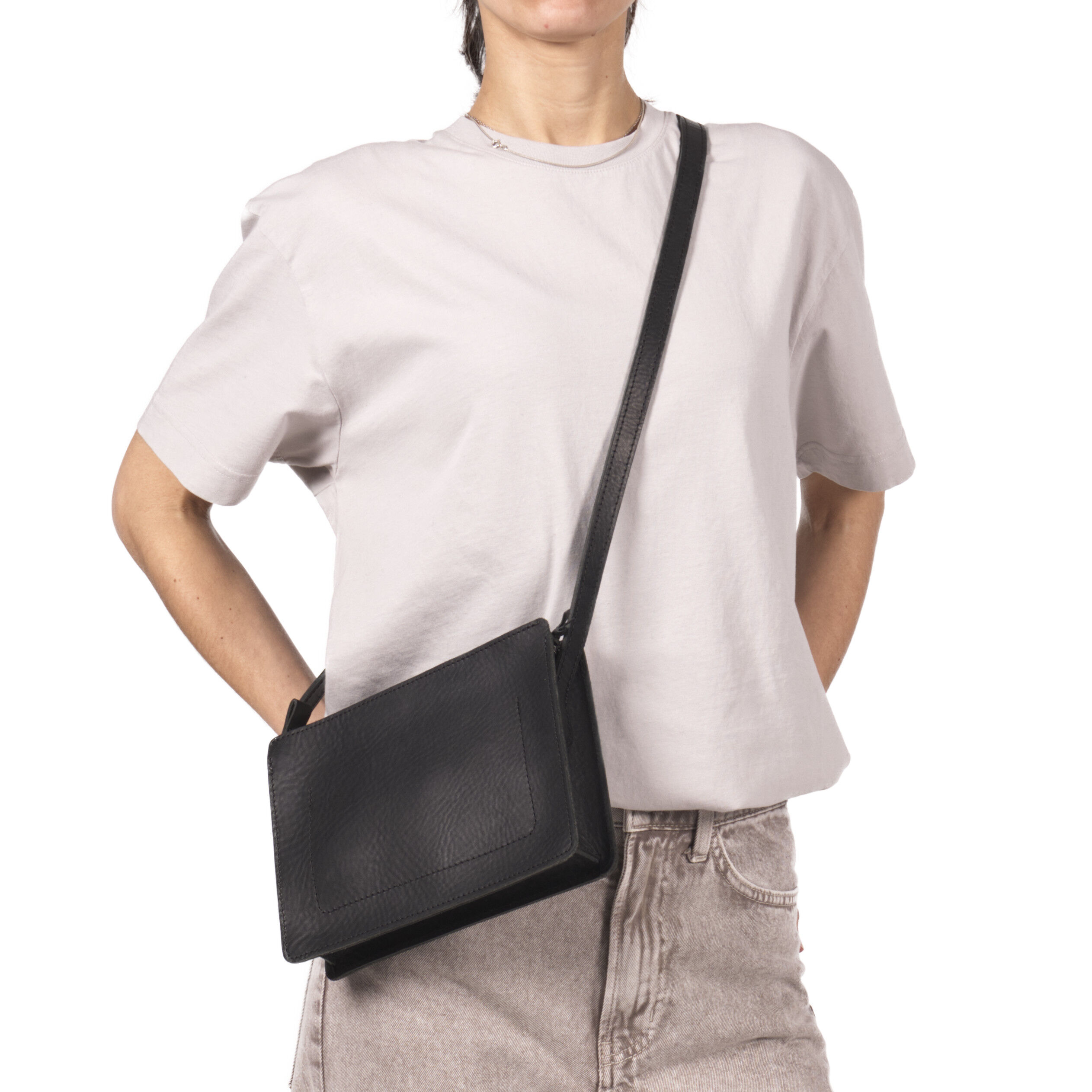 Eine Frau trägt eine schwarze Umhängetasche aus Kernleder mit schlichtem, rechteckigem Design. Die Tasche hat einen langen Schulterriemen und passt zu einem hellen T-Shirt und einer hellen Hose. Das stilvolle Accessoire aus Leder eignet sich für Alltag und besondere Anlässe.