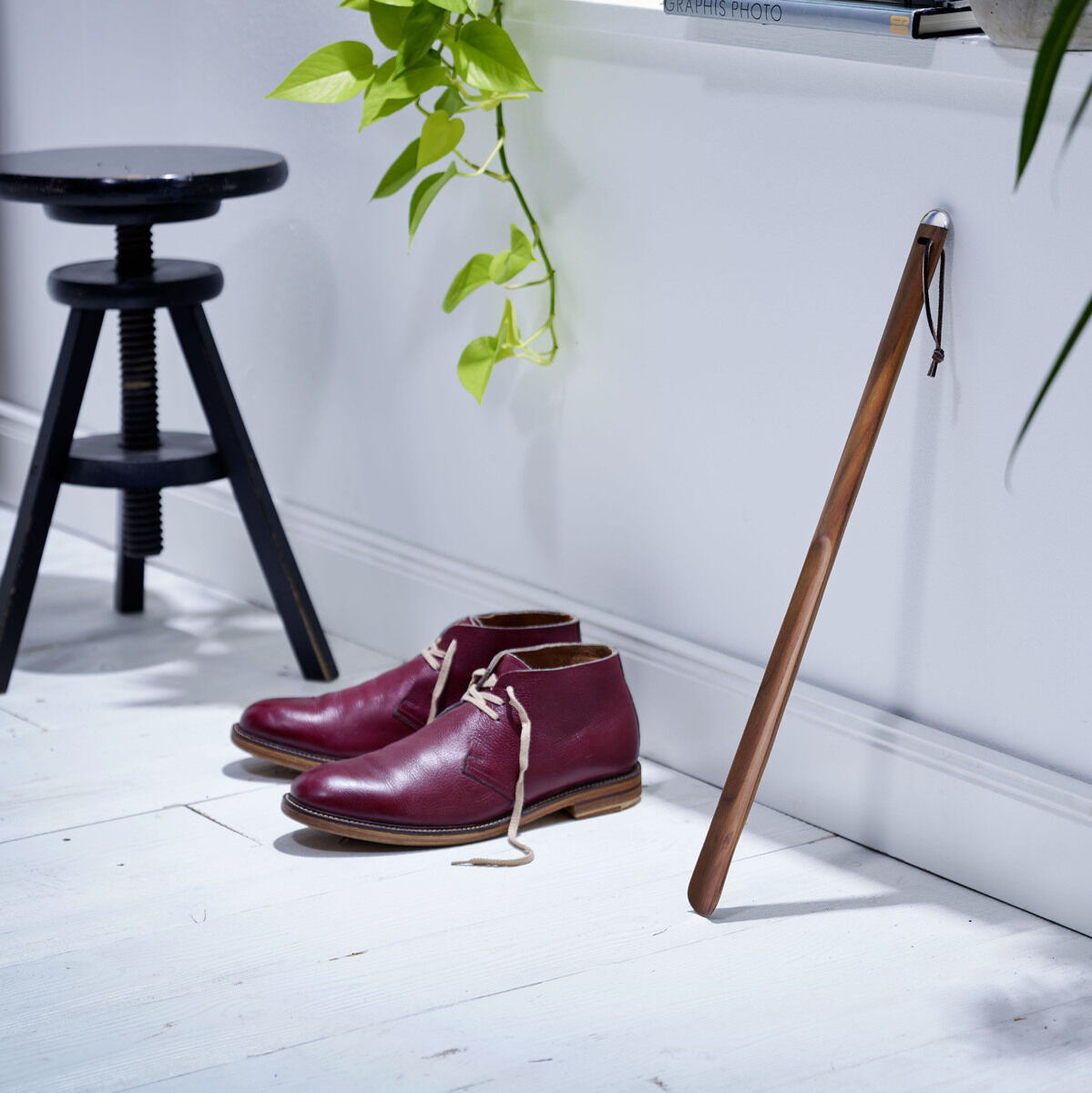 Holz-Schuhlöffel lehnt an einer weißen Wand, daneben ein Paar rote Schuhe auf weißem Boden, Pflanzen im Hintergrund.