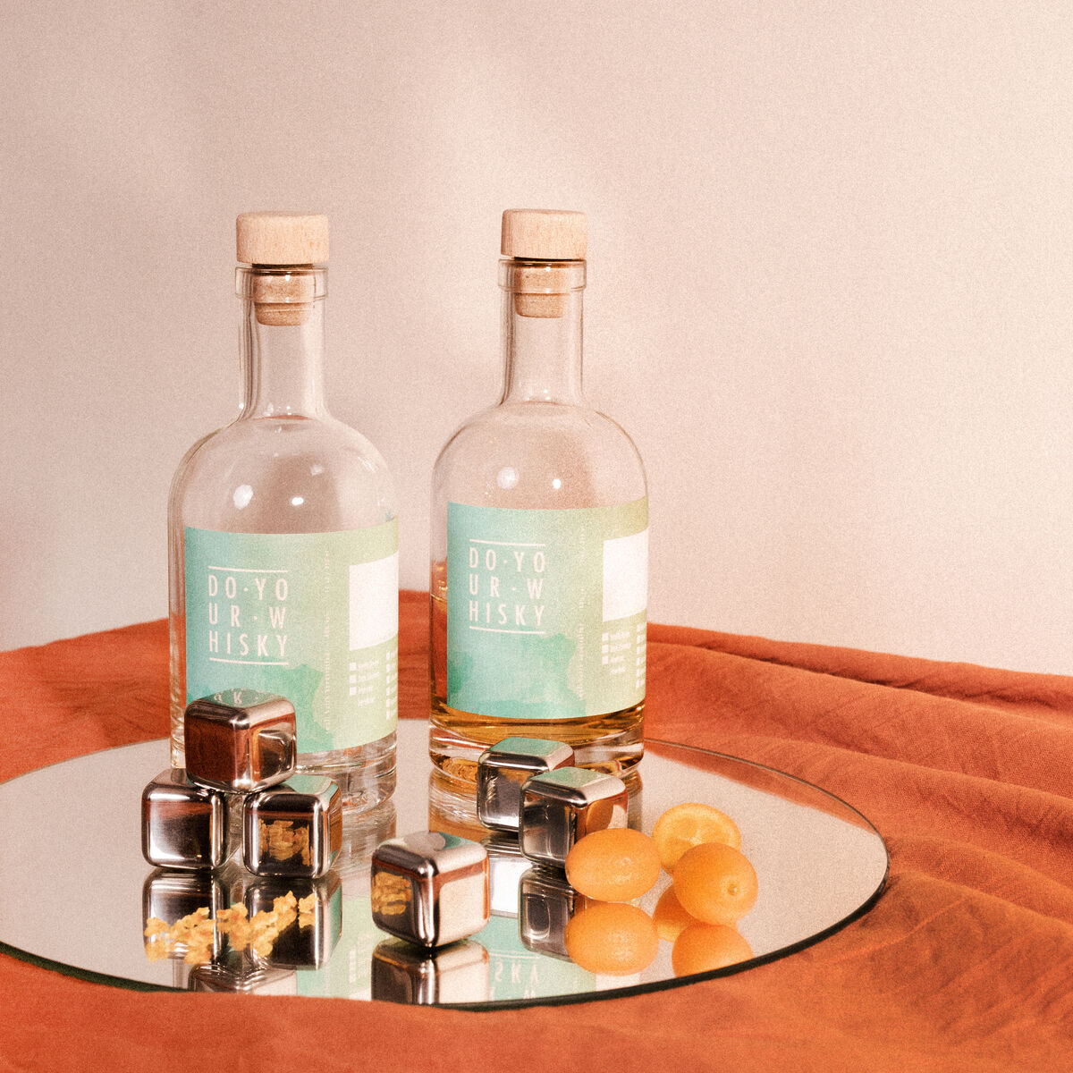 Ein Whisky-Set zum Selberherstellen, das zwei Glasflaschen mit Korkverschluss, Edelstahlkugeln und zwei gelbe Früchte enthält. Das Set ist auf einem silbernen Tablett vor einem orangefarbenen und beigen Hintergrund arrangiert.