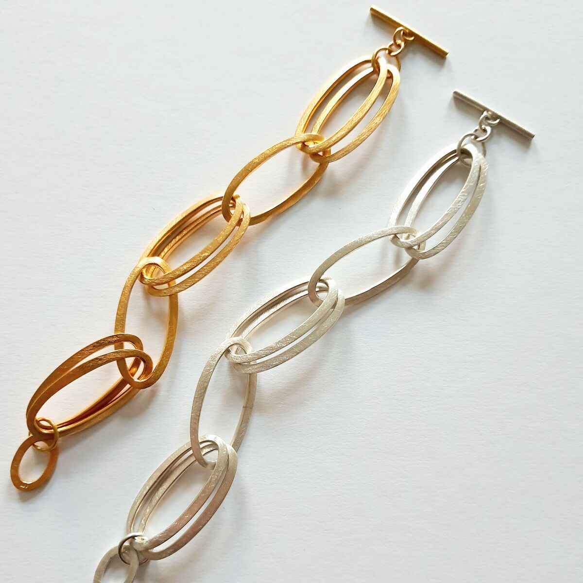 Zwei HML-Armbänder »GRACE« auf einer weißen Oberfläche. Ein Armband ist goldfarben, das andere silberfarben, beide bestehen aus ineinandergreifenden ovalen Gliedern.