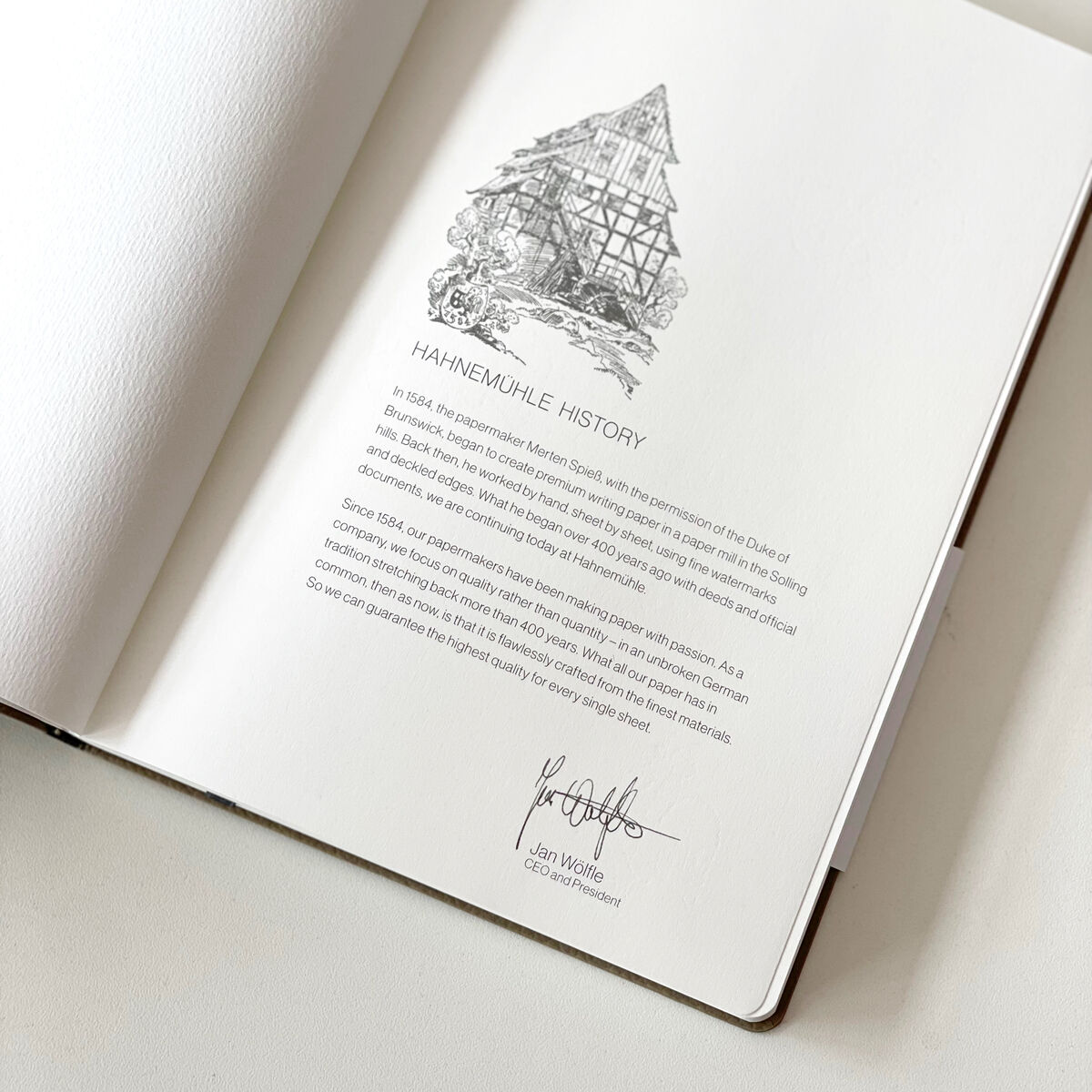 Ein geöffnetes Notizbuch »Iconic«, DIN A5, liegt auf einer weißen Oberfläche. Die dargestellte Seite zeigt eine handgezeichnete Illustration eines Hauses und den geschriebenen Text 'HAINEMUHLE HISTORY'.