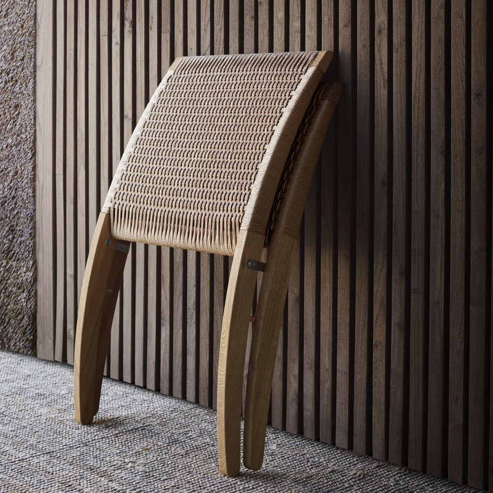 Der zusammengeklappte Stuhl »MG501 Cuba Chair« von Carl Hansen & Søn steht vor einer vertikal gerillten Holzverkleidung. Massivholzgestell in geölter Eiche, Sitzfläche aus Papiergarn-Geflecht. Schlichtes, elegantes Design für moderne Wohnräume.