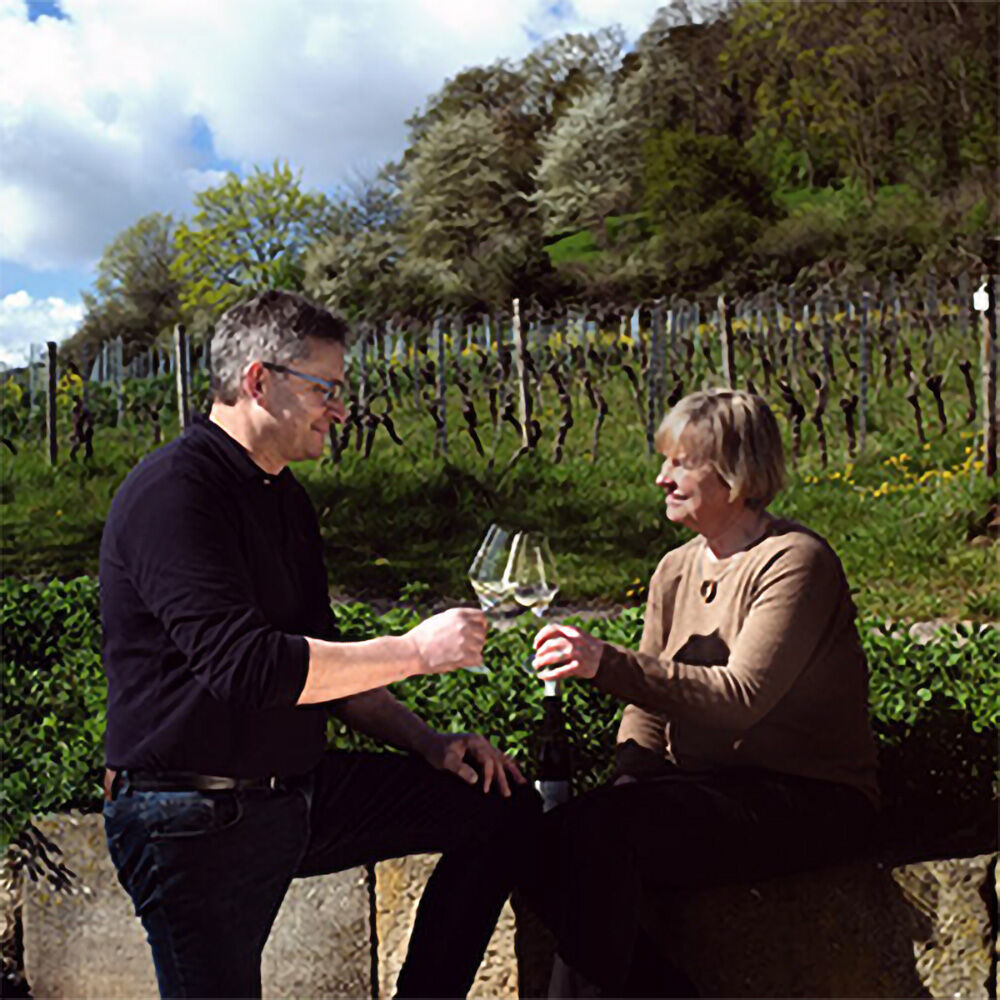 Ein Mann und eine Frau stoßen mit großen Weingläsern im Freien an. Sie sitzen auf einer niedrigen Steinmauer vor einem Weinberg mit blühenden Bäumen und Reben. Die Szene vermittelt Frühlingsatmosphäre und entspannten Weingenuss in der Pfalz.