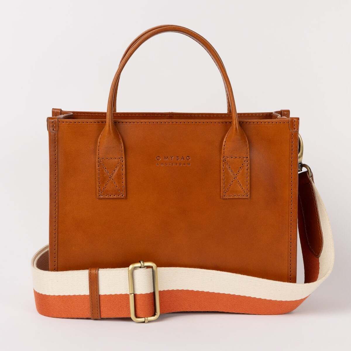 Rechteckige, mittelgroße Ledertasche in warmem Braunton mit kurzen Henkeln und abnehmbarem, cremeweiß-orangefarbenem Schulterriemen, stehend vor hellem Hintergrund.