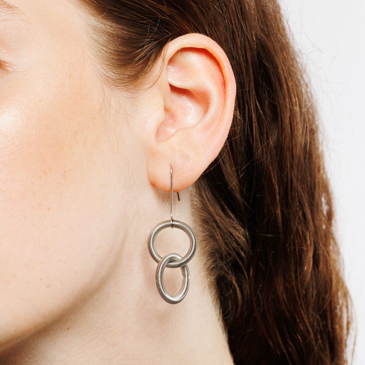 Detailaufnahme eines weiblichen Ohrs mit dem Ohrring "Oh Twin" von la molla aus zwei ineinanderhängenden silbernen Edelstahlfedern. Der Ohrschmuck wirkt filigran und modern, das Haar ist leicht gewellt und braun. Hintergrund neutral hellgrau.