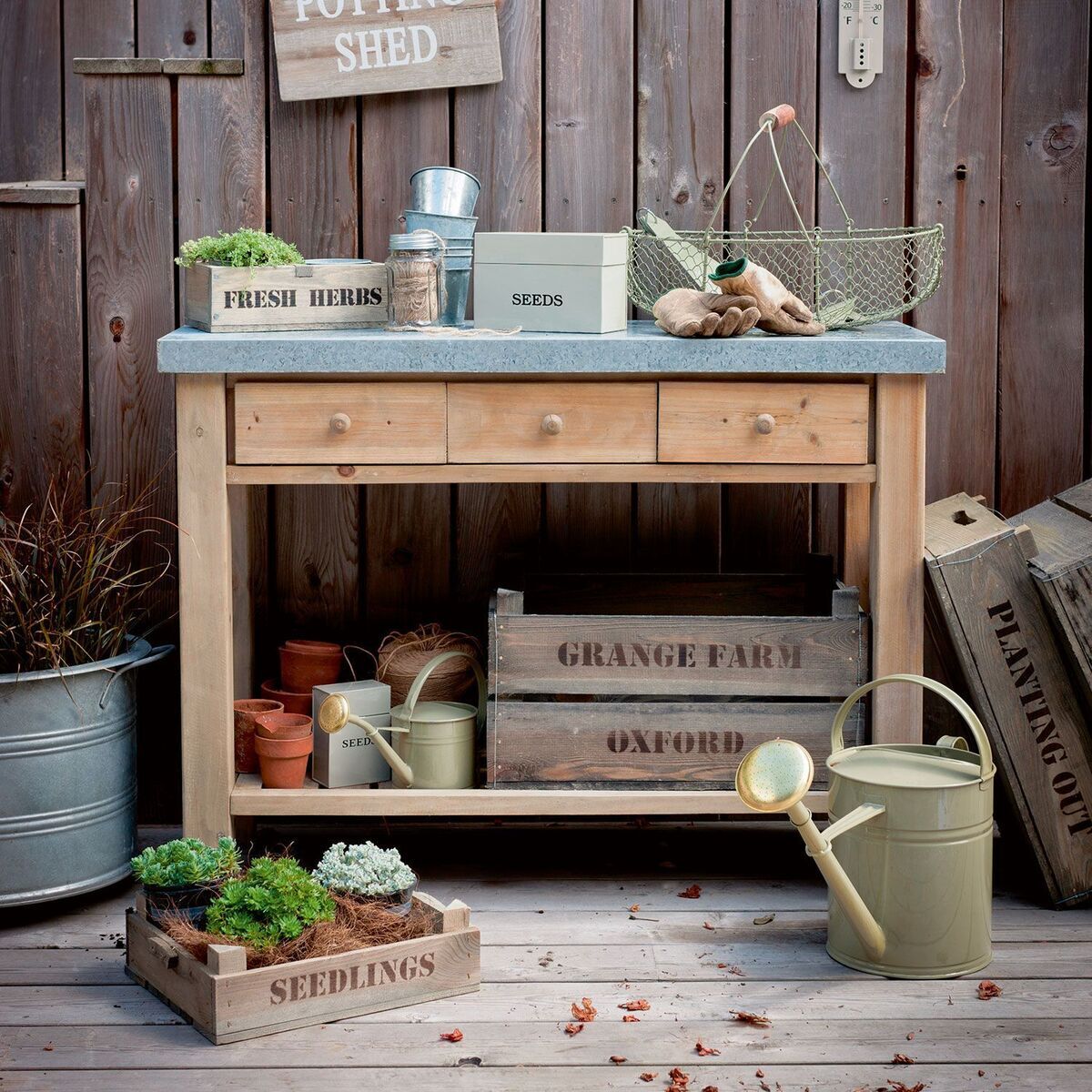 Ein rustikaler Garten-Arbeitstisch aus Holz, dekoriert mit verschiedenen Gartenwerkzeugen und Pflanzen. Der Tisch hat eine massive Arbeitsplatte und mehrere Schubladen sowie Ablageflächen. Rundherum befinden sich Pflanzboxen, Töpfe und ein Wasserkessel.