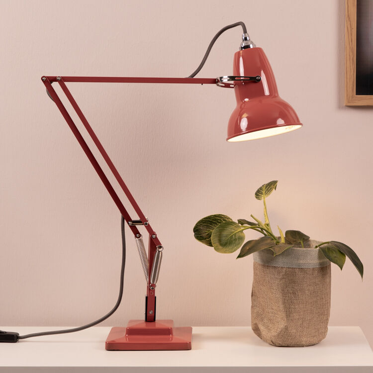 Eine rote Tischleuchte des Modells 'Anglepoise Original 1227' auf einem weißen Tisch. Neben der Leuchte befindet sich eine grüne Pflanze in einem beigen Topf. Der Hintergrund ist hellrosa, und ein gerahmtes Bild hängt an der Wand.