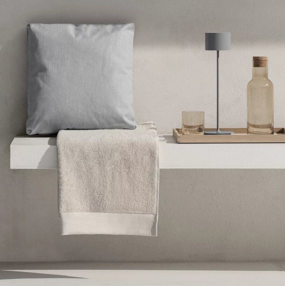 Mobile LED-Tischleuchte 'FAROL' auf einem minimalistischen Regal neben einem beigefarbenen Handtuch, einer grauen Kissenhülle und einer beigefarbenen Flasche. Das Design ist modern und schlicht, dominiert von neutralen Farben wie Grau, Beige und Weiß.