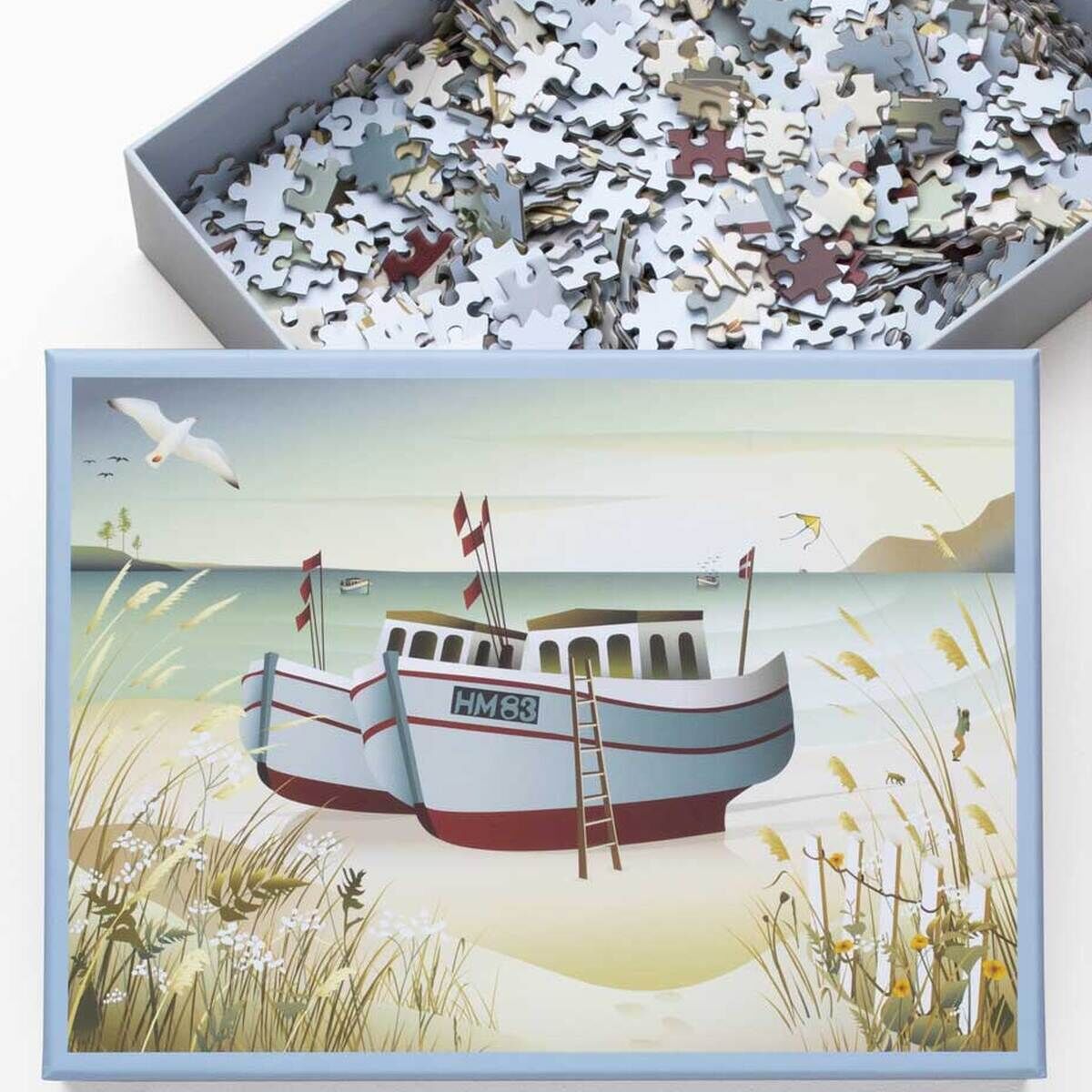 Ein Puzzle mit dem Motiv von Fischerbooten am Strand, umgeben von Meer, Sand, Gräsern und Möwen, das in einem Karton liegt.