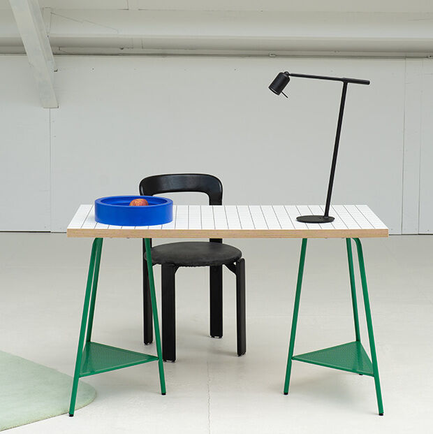 Ein minimalistischer Arbeitsplatz mit einem Tisch, auf dem eine schwarze Schreibtischlampe der Marke »ONE« mit Halterung steht. Rechts neben dem Tisch steht ein schwarzer Stuhl. Der Tisch hat grüne Beine und auf der Tischplatte befindet sich eine blaue Schale.