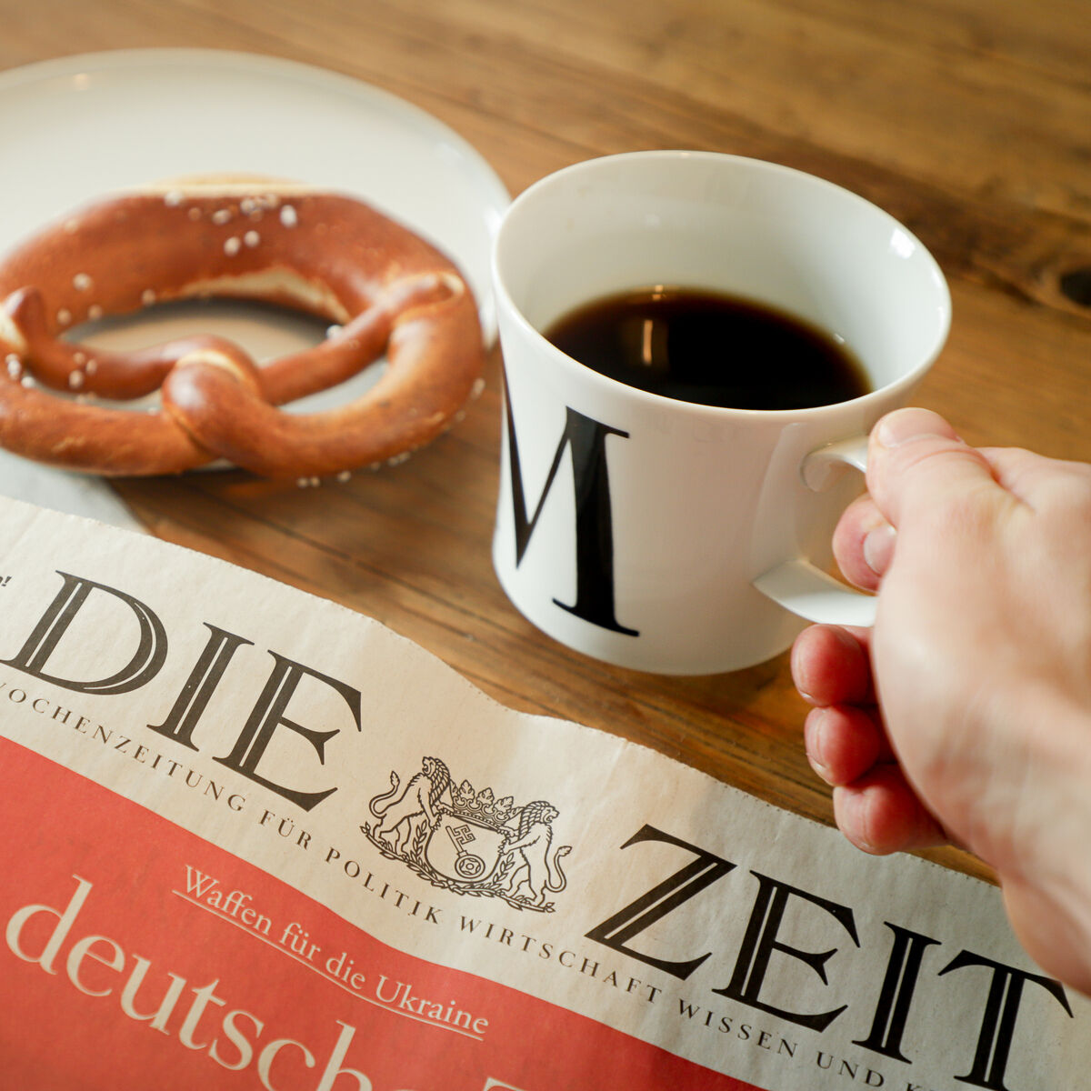Eine weiße Frühstückstasse mit dem Buchstaben 'M' darauf, gefüllt mit Kaffee. Daneben liegt eine Brezel auf einem weißen Teller und eine Zeitung mit dem Titel 'DIE ZEIT' auf dem Tisch. Eine Hand hält die Tasse. Das Bild vermittelt eine gemütliche Morgenstimmung beim Frühstücklesen.