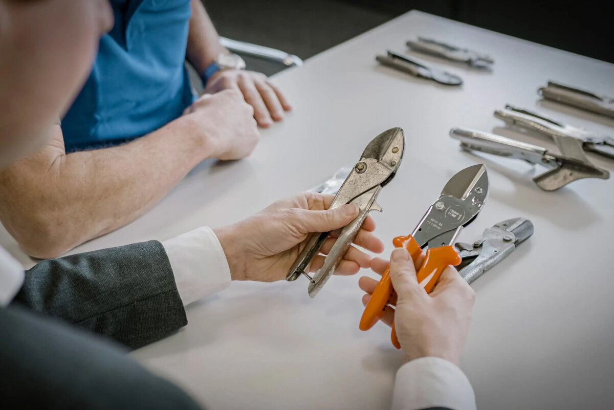Hände halten zwei verschiedene Zangen, eine mit Metallgriffen, die andere mit orangefarbenen Kunststoffgriffen, auf einem Tisch mit weiteren Werkzeugen.