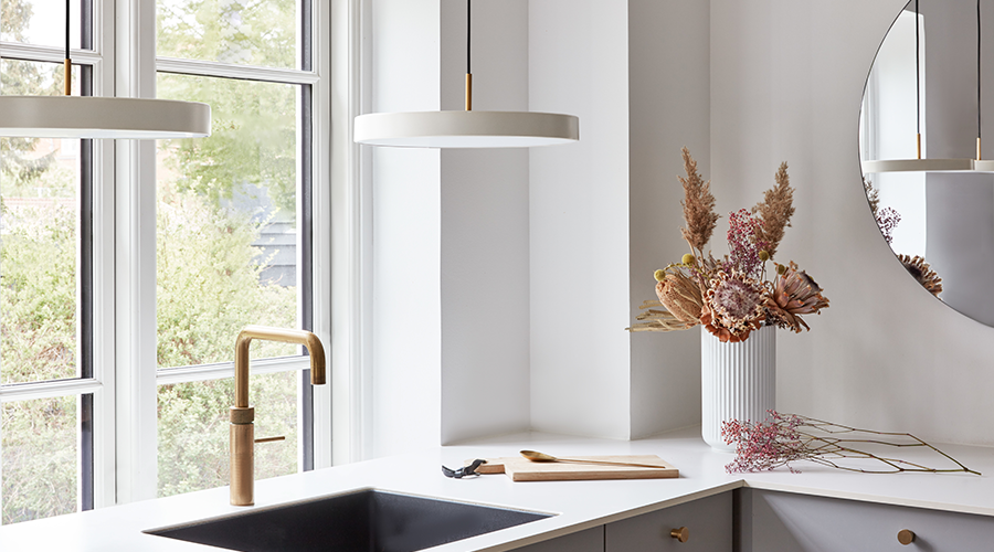 Moderne Küche mit schwarzer Spüle, goldfarbenem Wasserhahn, weißen Arbeitsflächen und Trockenblumen in weißer Vase neben rundem Spiegel