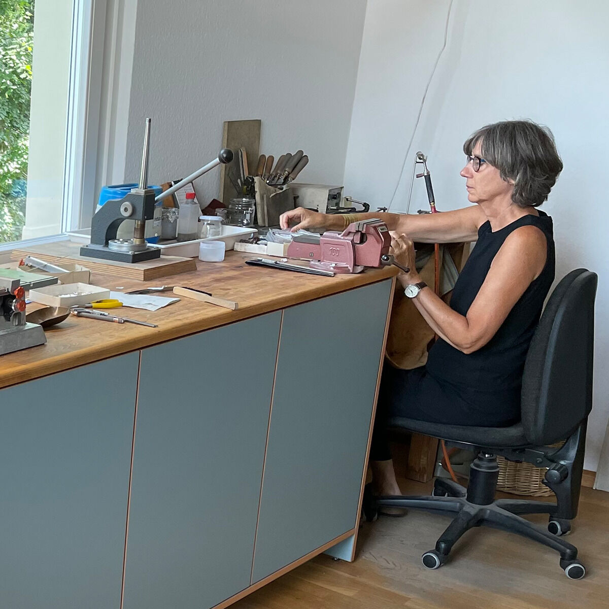 Eine Frau sitzt in heller Werkstatt an Holzarbeitsplatte, umgeben von Werkzeugen, Schmuckelementen und Werkmaterialien. Sie arbeitet konzentriert an einem Schmuckstück und trägt ein schwarzes ärmelloses Outfit. Moderne, minimalistische Einrichtung mit großem Fenster im Hintergrund.