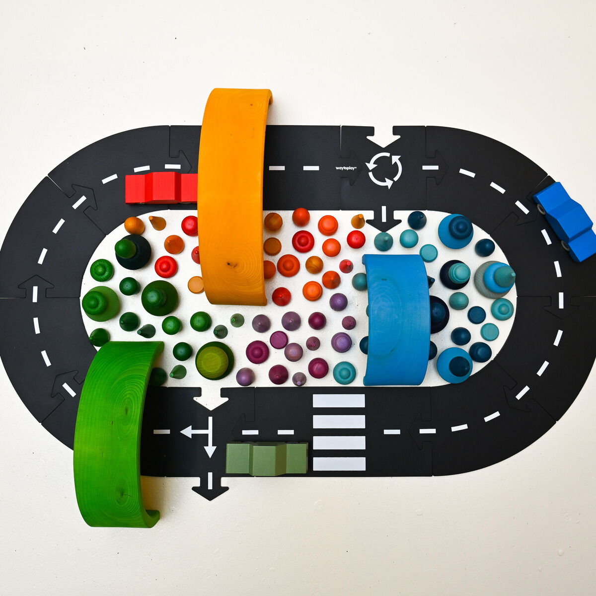 Das Bild zeigt ein Straßenbau-Set mit einer ovalen schwarzen Fahrbahn inklusive Markierungen, Zebrastreifen und Verkehrszeichen. Innerhalb der Strecke befinden sich bunte Holzzylinder und -kugeln in verschiedenen Farben sowie zwei große bunte Brücken. Zwei kleine Fahrzeuge, rot und blau, sind auf der Straße platziert. Das Spielzeug liegt auf einer hellen Unterlage und ist für Kinder geeignet.