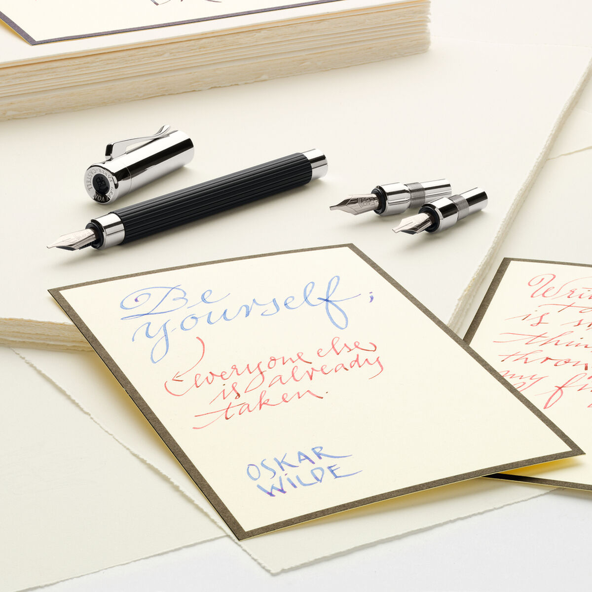 Auf dem Bild ist das Tamitio Kalligraphie Set zu sehen, bestehend aus einem schwarzen Füllfederhalter mit mehreren austauschbaren Federn. Das Set liegt auf cremefarbenem Briefpapier, auf dem handschriftlich kalligrafierte Karten in blauer und roter Tinte zu sehen sind. Daneben befinden sich weitere Papierstapel, das Bild vermittelt eine elegante und kreative Atmosphäre.