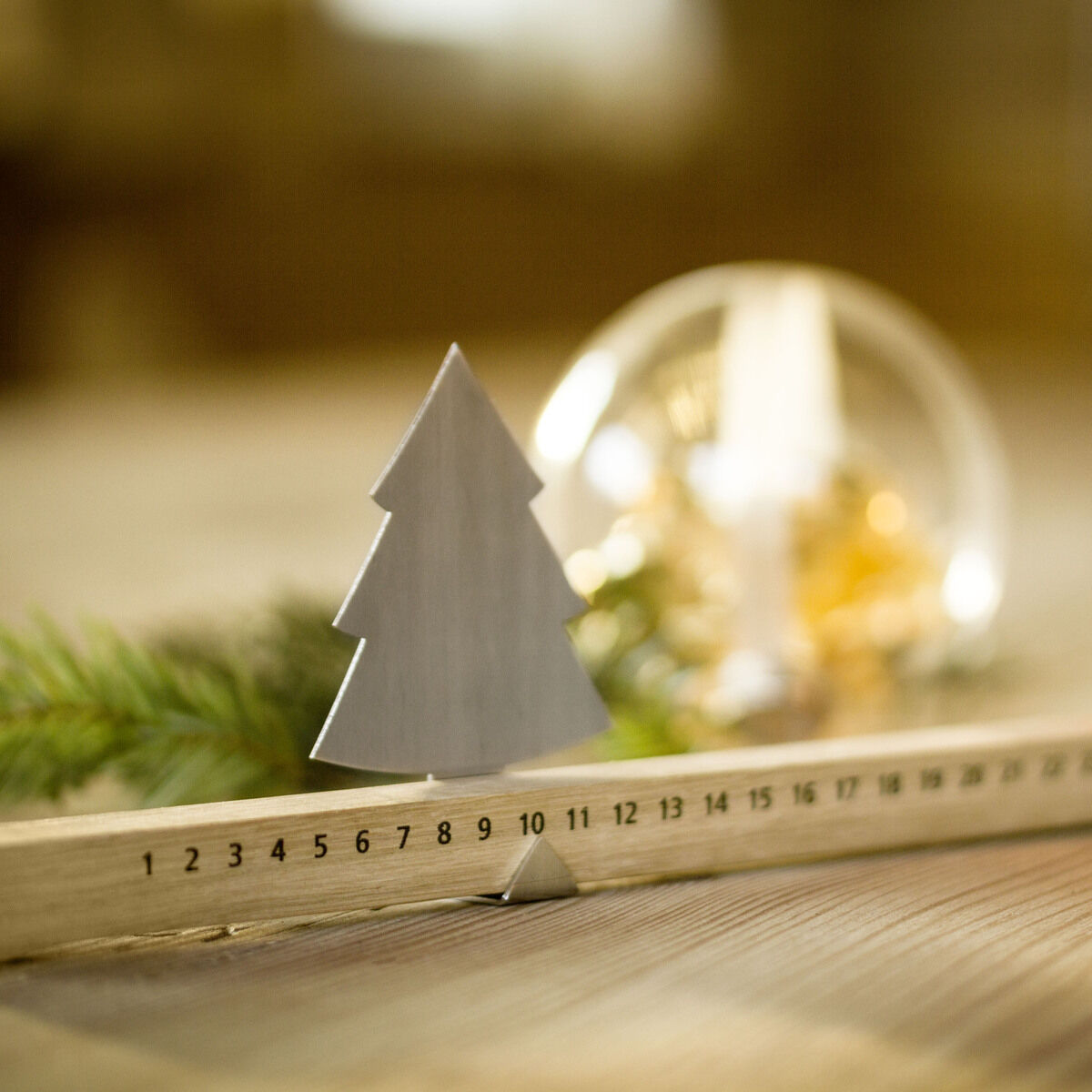 Ein Adventskalender in Form einer Holzleiste mit nummerierten Tagen bis Weihnachten, verziert mit einem kleinen Tannenbaum und einem brennenden Teelicht. Im Hintergrund befinden sich weihnachtliche Dekorationen und grüne Zweige.
