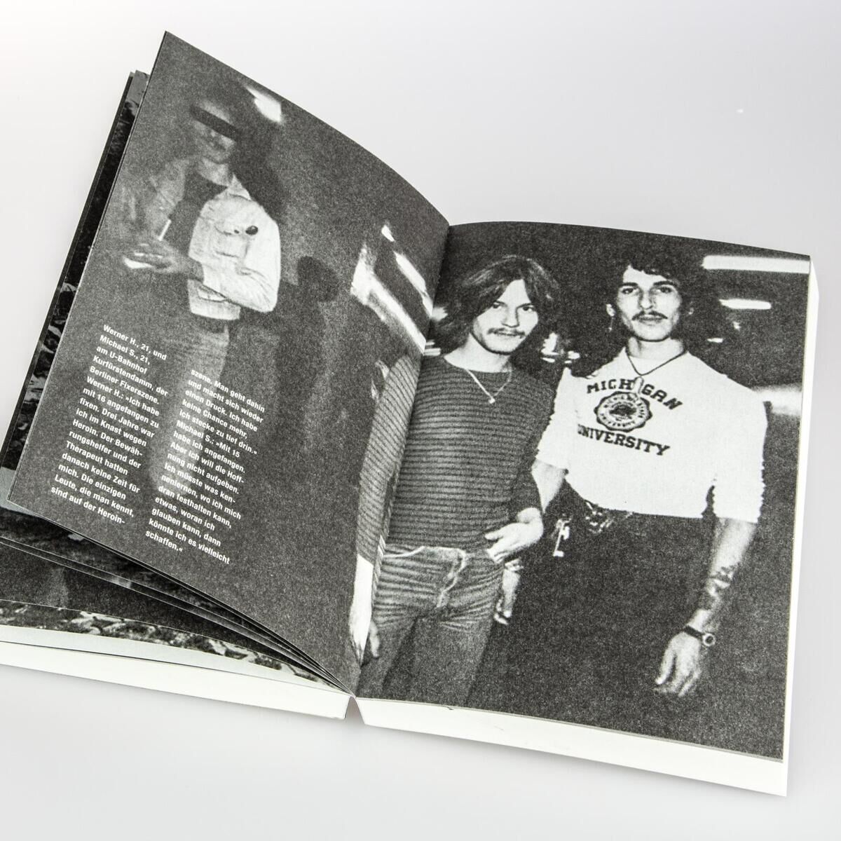 Eine aufgeschlagene Seite des Buches 'Wir Kinder vom Bahnhof Zoo', die ein Schwarz-Weiß-Foto von zwei jungen Menschen zeigt. Einer trägt ein langärmliges Shirt, der andere ein T-Shirt mit der Aufschrift 'Michigan University'.