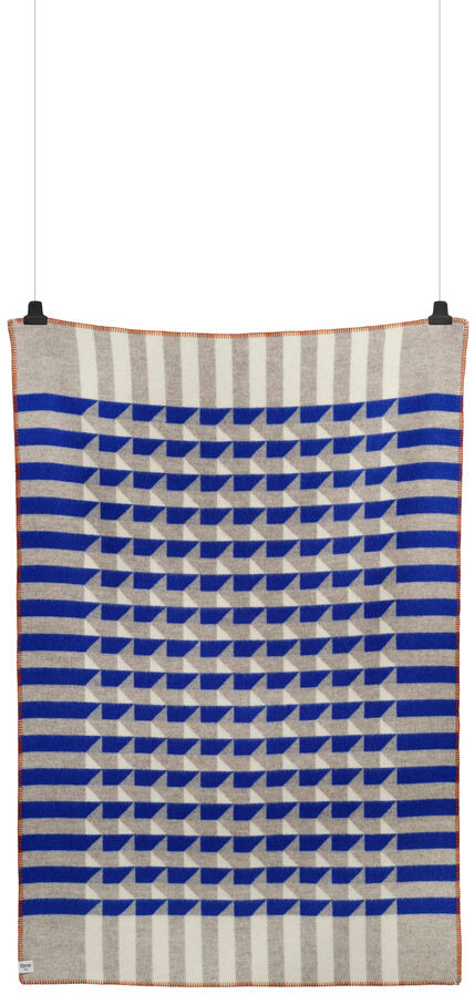 Eine Lammwolldecke mit dem Namen »KVAM« zeigt ein geometrisches Design mit blauen, beigen und braunen Streifen. Die Decke ist aufgehängt und wurde für luxuriöse Gemütlichkeit in modernen Wohnräumen entworfen.