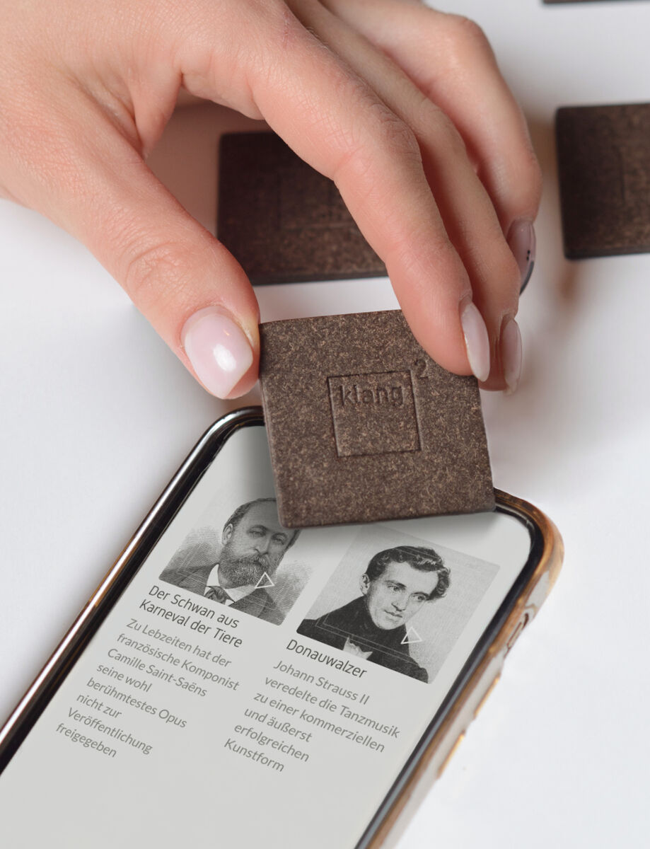 Eine Hand hält ein rechteckiges Holzstück mit der Aufschrift „klang²“ über ein Smartphone, auf dessen Bildschirm zwei Porträts und Texte von historischen Persönlichkeiten sichtbar sind.
