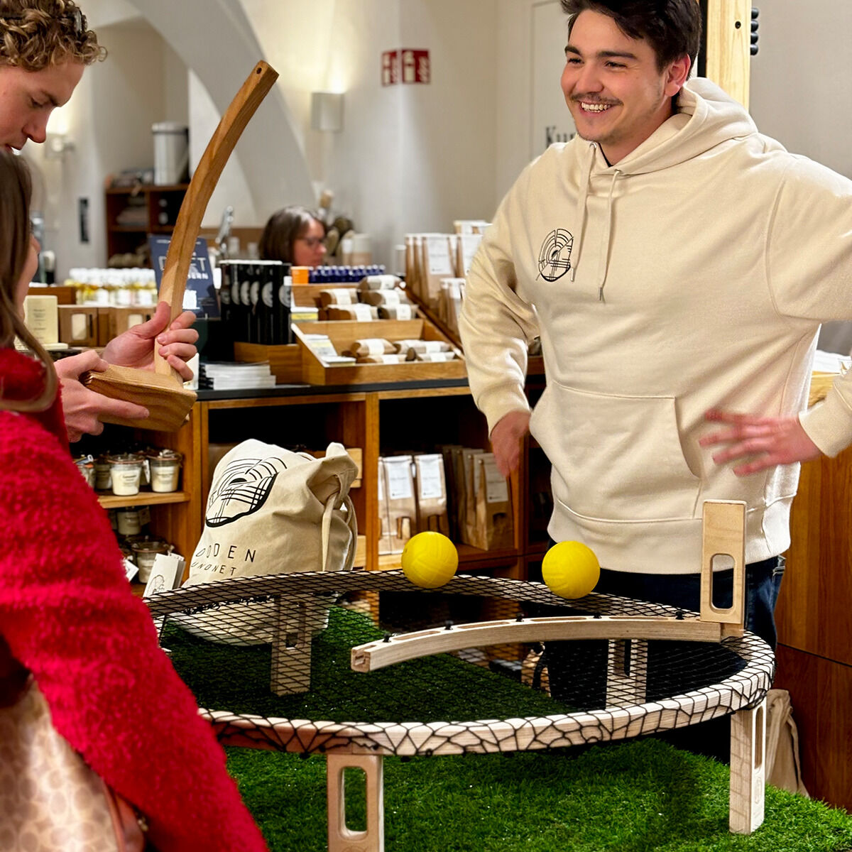 Personen spielen im Innenraum zusammen ein Spiel namens Wooden Roundnet. Es gibt ein rundes Netz auf einem Tisch mit gelben Bällen darauf. Eine Person hält ein hölzernes Zubehör. Im Hintergrund sind Regale und Bücher zu sehen. Die Stimmung ist entspannt und fröhlich.
