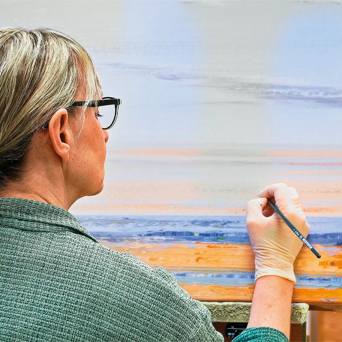 Eine Frau, die ein Gemälde einer Meereslandschaft malt, mit sanften Blautönen, Weißen und Orangen Farbakzenten, Titel: 'Leises Meeresrauschen' von Anja Struck aus dem Jahr 2022.