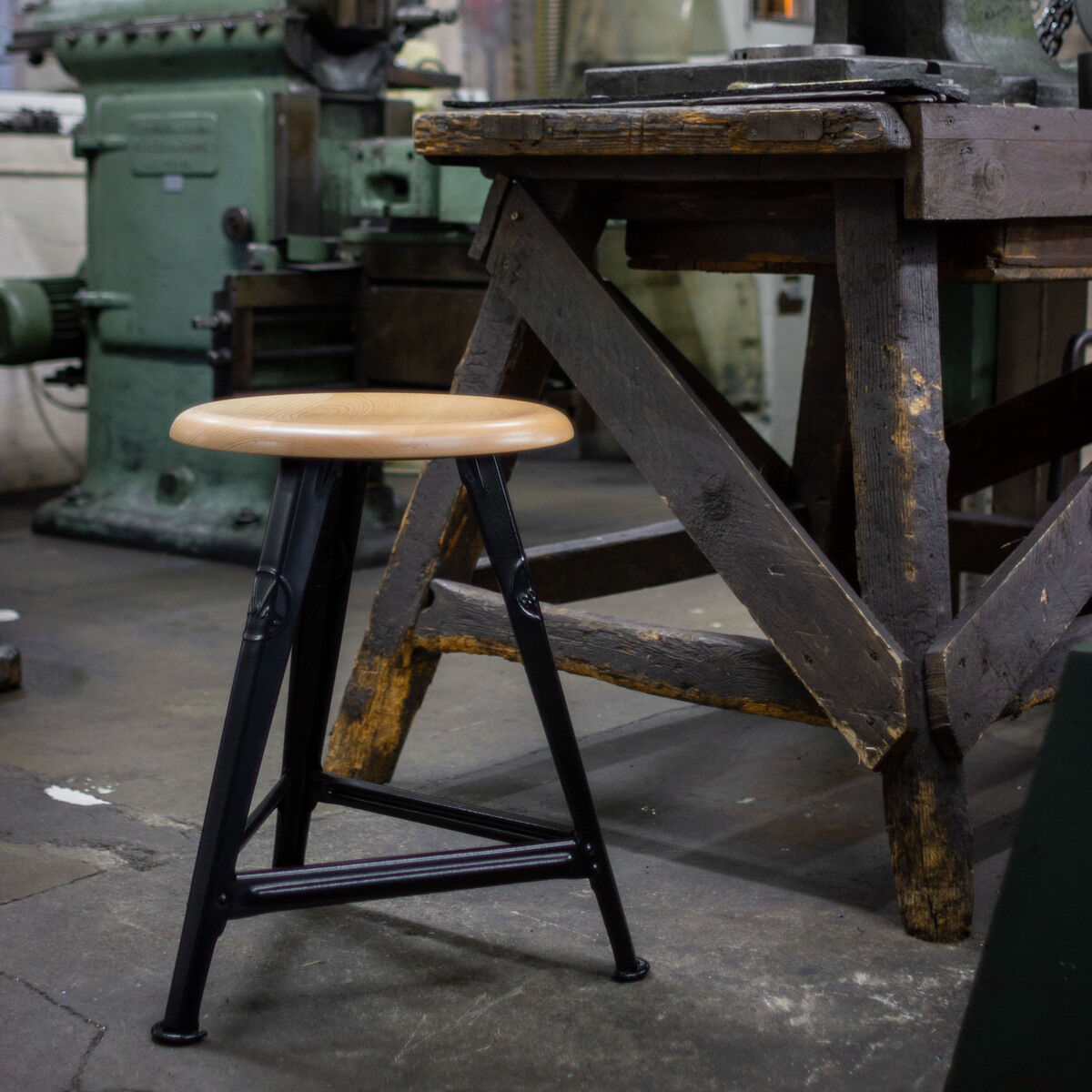 Ein industrieller Hocker mit einer eichenfarbenen Sitzfläche und einem mattschwarzen Metallgestell steht in einer rustikalen Werkstatt neben einem alten Holztisch und großen Maschinen.