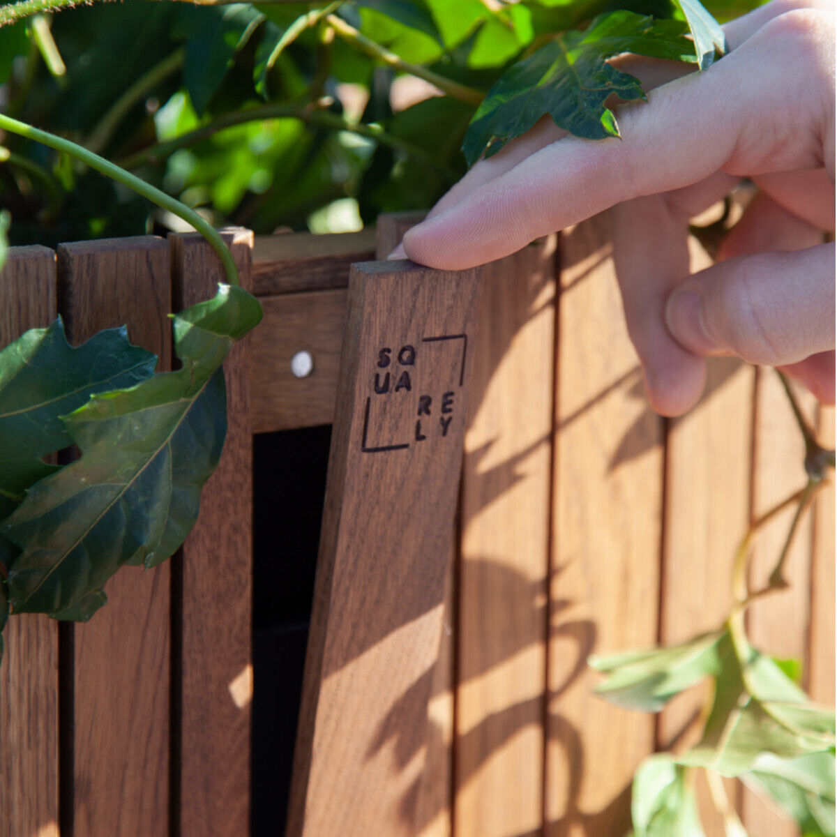 Ein handgefertigtes, quadratisches Pflanzgefäß aus Holz mit grünen Pflanzen, das von einer Hand berührt wird. Perfekt für Urban Gardening und nachhaltige Gartenarbeit.