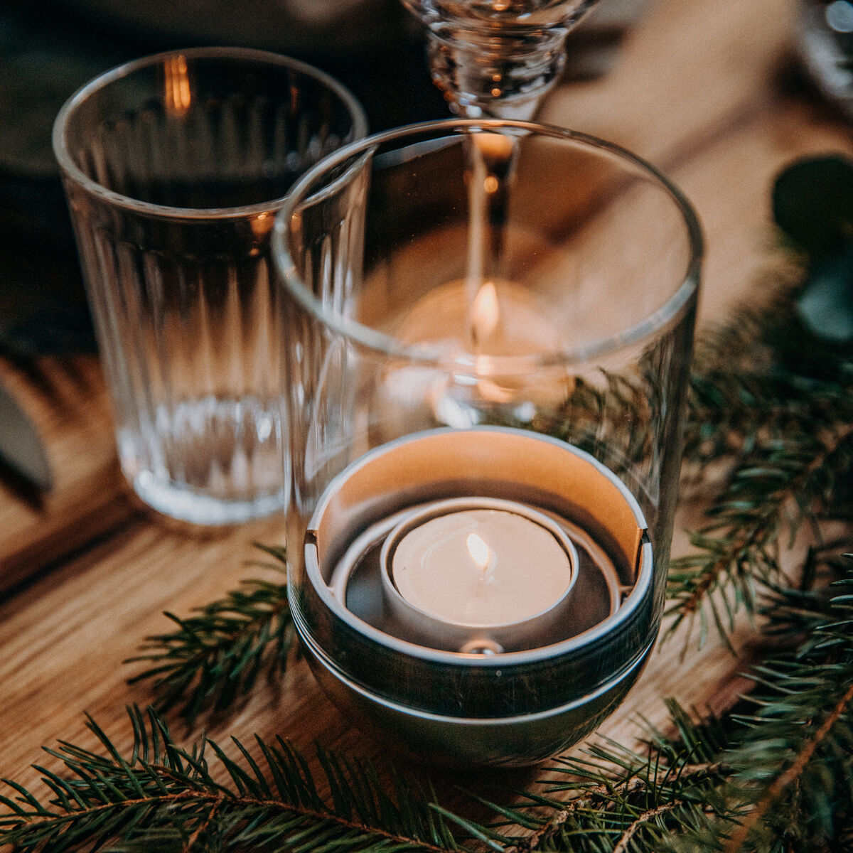 Eine stimmungsvolle Tischdekoration mit einer Gravity Candle in einem transparenten Glas, umgeben von grünen Tannenzweigen auf einem Holztisch. Die Flamme der Kerze brennt ruhig und schafft eine gemütliche Atmosphäre.