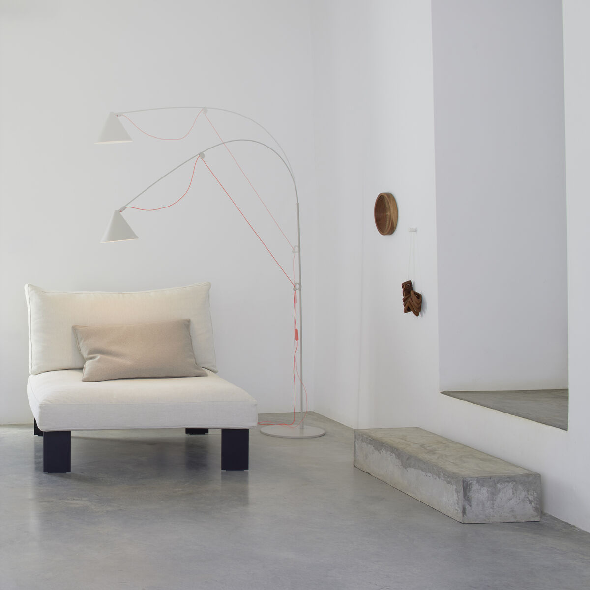 Eine weiße Stehlampe AYNO Silk mit minimalistischer und verstellbarer Struktur steht neben einem weißen Sofa mit hellbeigem Kissen in einem modern gestalteten Wohnraum mit Betonboden und weißen Wänden.