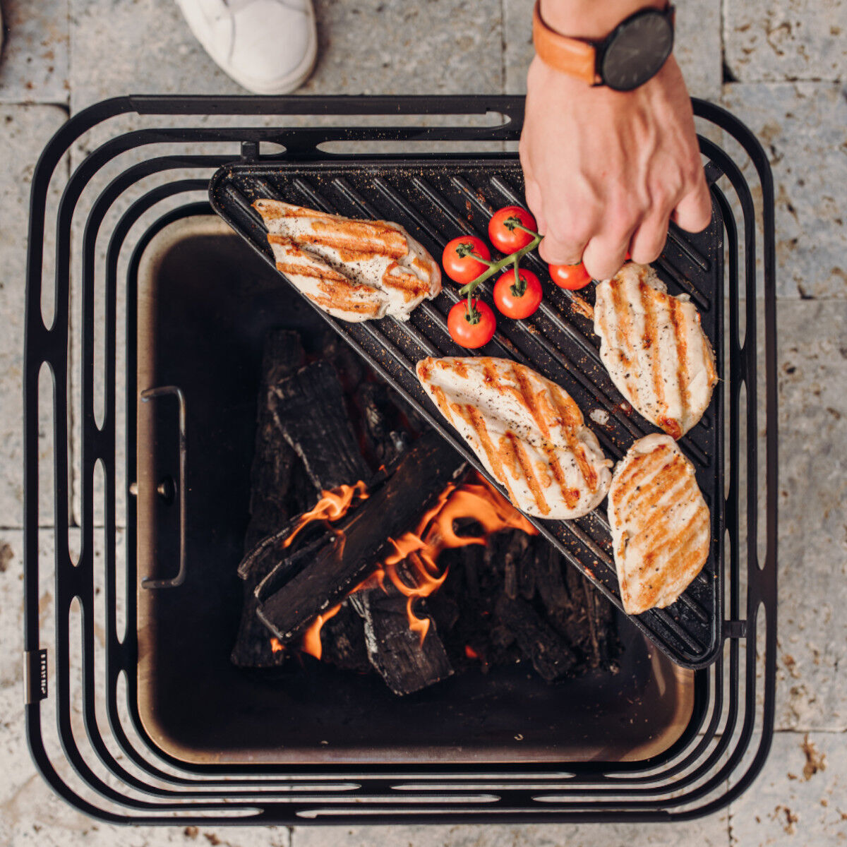 Eine »CUBE« Plancha mit gegrillten Hähnchenfilets und Cherry-Tomaten auf einem Grillrost über offenen Flammen und glühender Holzkohle. Eine Hand wendet das Fleisch. Der Hintergrund zeigt eine helle Steinfläche.